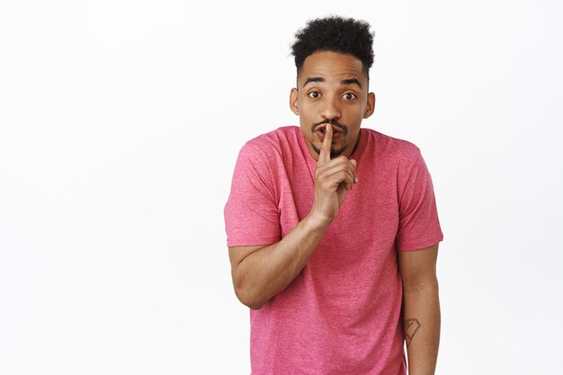 Hush its secret. Portrait of young african american guy shushing, press finger to lips and prepare surprise, shhh gesture, hiding smth, standing against white background in blue t-shirt