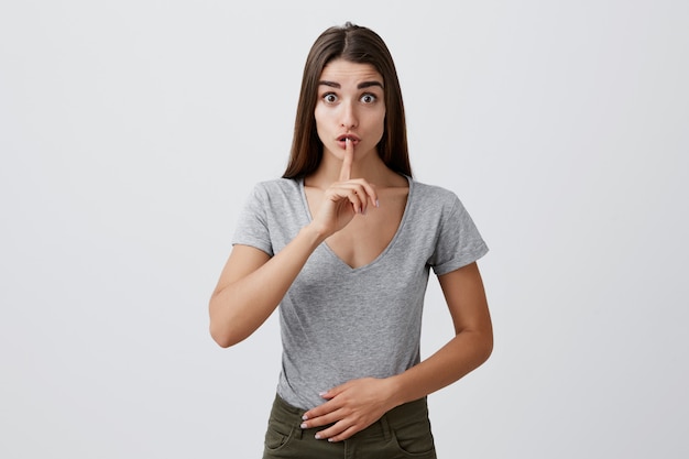 Hush please. Portrait of young attractive charming caucasian woman with dark long hair in casual stylish clothes holding index finger in front of lips with raised eyebrows.