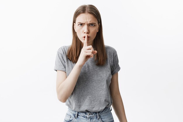 Hush. Don't scream. Attractive young brunette student girl in grey t-shirt holding finger near lips,  with unsatisfied face expression trying to calm down shouting child in bus.