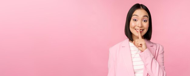 Hush taboo concept Portrait of asian businesswoman showing shush gesture shhh sign press finger to lips standing over pink background in suit