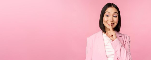 ハッシュタブーの概念スーツのピンクの背景の上に立っている唇にshushジェスチャーshhhサインプレス指を示すアジアの実業家の肖像画