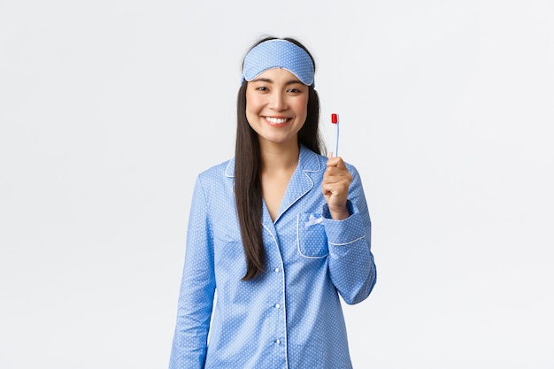 Гигиена, образ жизни и люди дома концепции. Веселая улыбающаяся азиатская девушка в пижаме и спальной маске, показывающая зубную щетку и белые идеальные зубы, использует отбеливающую зубную пасту, белый фон.