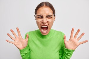 Free photo hysterical mad european woman feels crazy keeps palms raised screams loudly from anger wears green jumper and big transparent glasses isolated over white background. negative emotions concept