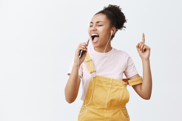 I am best in karaoke. Portrait of carefree excited and attractive female with dark skin in yellow overalls, listening music and singing along in earphones, making microphone from smartphone