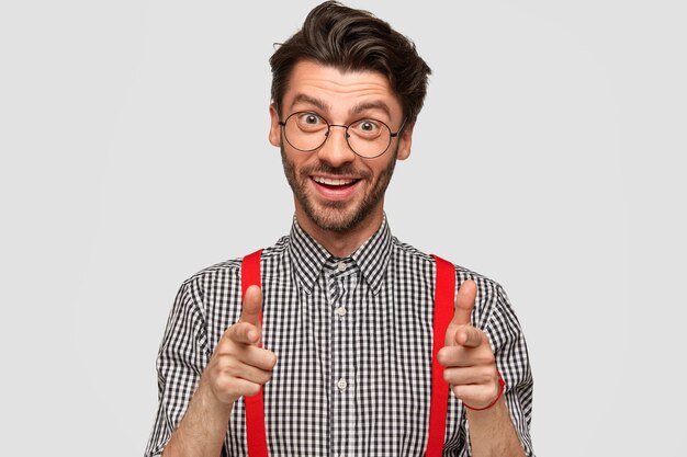 I choose you! Smiling positive young male businessman points directly with both index fingers, expresses his choice, has happy expression, dressed in checkered shirt and red braces