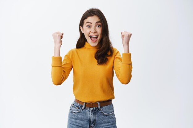 I did it. Excited girl rejoicing, raising hands up and smiling happy, achieve goal or prize, winning money and celebrating, standing in casual clothes on white background