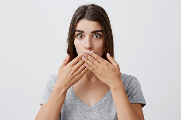 I didn't say that. Beautiful young caucasian brunette girl with long hairstyle in fashionable casual gray t-shirt clothing mouth with hands,  with confused and surprised look.