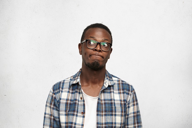 I don’t know. Clueless funny young dark-skinned male wearing stylish glasses pursuing lips and looking sideways with doubtful and uncertain expression, feeling indecisive about his future plans