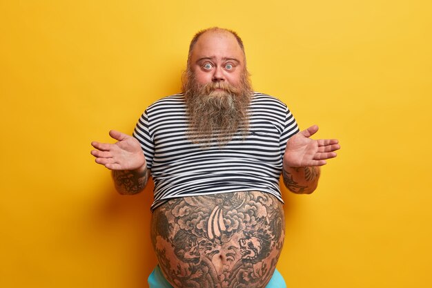 I dont care about rules. Indifferent careless questioned bearded man shrugs shouldres with hesitation, puzzled by stupid question, has very big tummy, wears striped t shirt, no idea how it happened