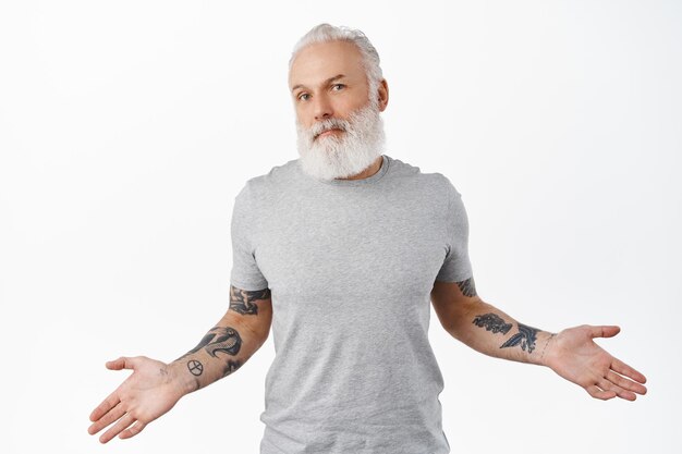 I dont know. clueless senior man with tattoos spread hands sideways, shrugging and looking confused, have no idea, being indecisive and unaware, white background.