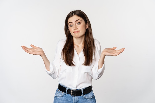 I dont know. Clueless young woman shrugging shoulders and looking unaware, standing over white background. Copy space