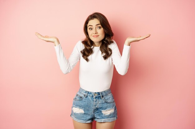 I dont know. Confused cute young woman smiling awkward, shrugging shoulders clueless, has no idea, standing over pink background