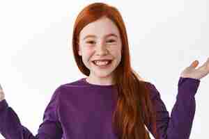 Free photo i dont know. happy smiling ginger girl spread hands sideways in unaware and clueless gesture, have no idea, standing careless and unbothered against white background