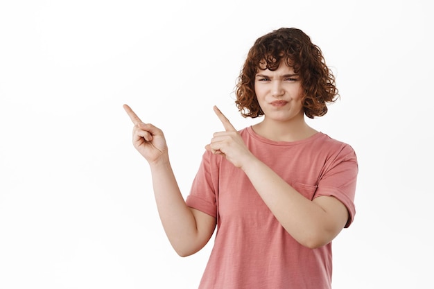 I dont think so. Reluctant and doubtful cute girl, grimacing and frowning from dislike, pointing at something bad, upper left corner, standing in t-shirt against white background