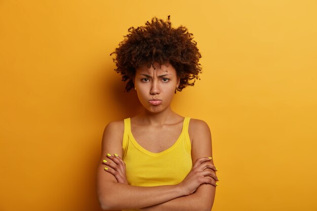 I dont trust you. Gloomy dissatisfied woman keeps arms folded, looks with miserable expression  from under forehead, dressed casually, poses over yellow wall, makes unhappy grimace