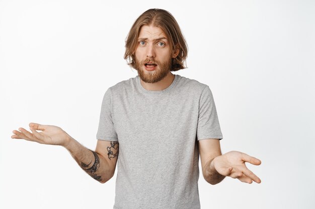 I dont understand. Confused and clueless blond guy shrugging shoulders, looking puzzled, know nothing, being unaware, standing in grey t-shirt on white.