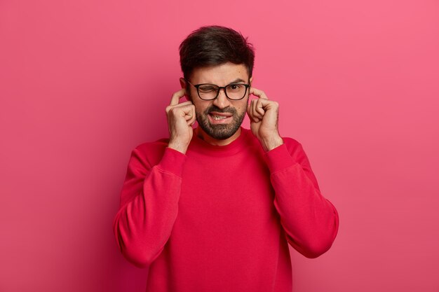 I dont want to listen it! Annoyed unshaven man winks eye, hears loud noise, avoids unpleasant sound, wears glasses and red jumper, doesnt hear somebodys advice, closes ears with index fingers