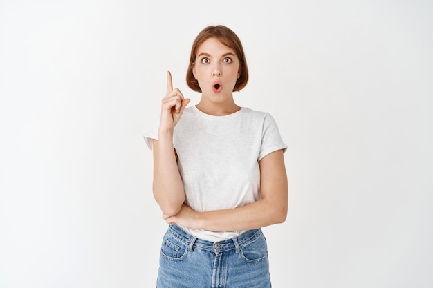 I got idea. Excited young woman in t-shirt, raising finger eureka sign, have plan, suggesting something, standing on white wall