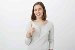 Free photo i got your back covered. portrait of friendly loyal and optimistic attractive woman with short brown haircut winking and smiling joyfully showing thumb up in like and supportive gesture over gray wall