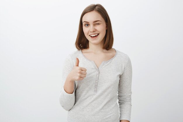 I got your back covered. Portrait of friendly loyal and optimistic attractive woman with short brown haircut winking and smiling joyfully showing thumb up in like and supportive gesture over gray wall