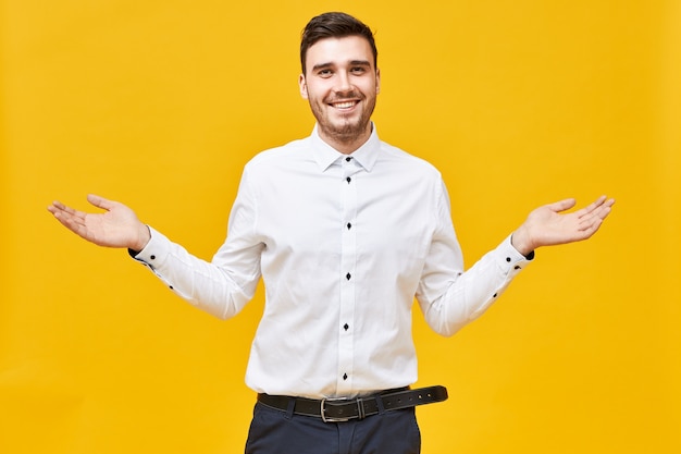 I have no idea.  emotional attractive young unshaven man smiling happily, spreading arms wide, making welcoming gesture, showing presentation, advertising product at copyspace wall