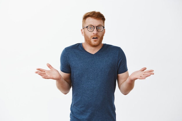 I have no idea how that happened. Portrait of clueless cute masculine male with red hair in glasses and t-shirt, shrugging with spread palms and gazing confused and uncertain over grey wall