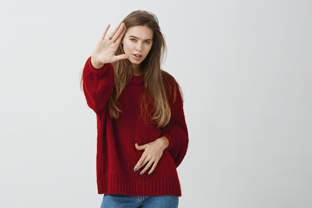 あなたを止める力があります。真剣で自信を持っている、停止または十分なジェスチャーでカメラに向かって手のひらを引っ張るルーズな赤いセーターの魅力的な集中少女のスタジオ撮影。