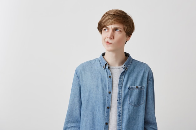 I don't know. Doubtful caucasian young male wearing denim shirt, pouting lips and looking up with indecisive expression on his face, showing doubt and hesitation. Body language and face expression