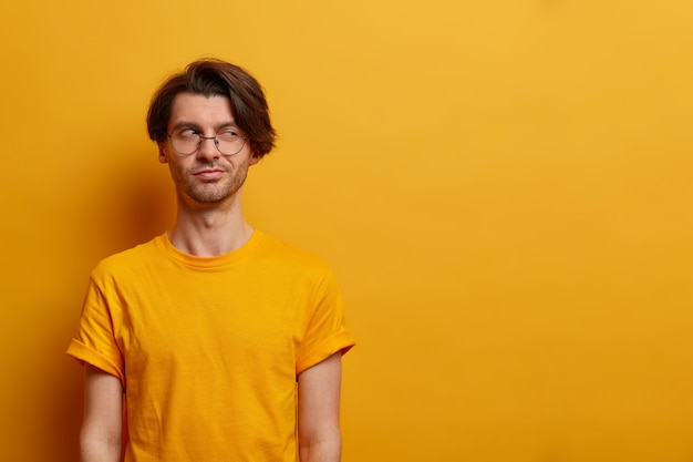 Free photo i know what i need. devious mysterious adult man looks with sly expression aside, has evil intention, nice idea or scheme, poses against yellow wall, empty space