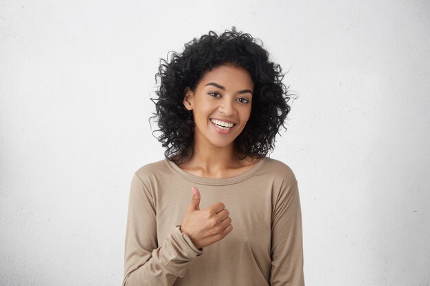 私はすきです。よくやった。幸せな若い暗い肌の女性がカジュアルな長袖Tシャツを着て親指を立て、元気に笑って、誰かへの彼女のサポートと敬意を示しています。ボディランゲージ