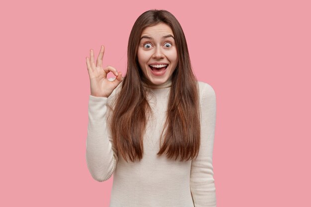 I like when everything is perfect. Delighted European woman shows okay gesture, smiles broadly, dressed in white turtleneck sweater