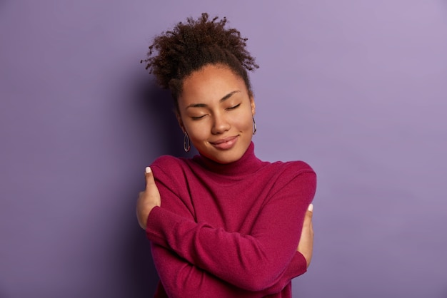 Free photo i love myself. tender romantic woman embraces own body, hugs herself, closes eyes from pleasure, wears soft turtleneck just for cold weather, feels comfort, stands indoor against purple wall