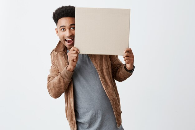 I'm here. Positive emotions. Copy space for advertisement. Close up of young good-looking afro american with curly dark hair in casual autumn clothes looking out in camera from carton board.