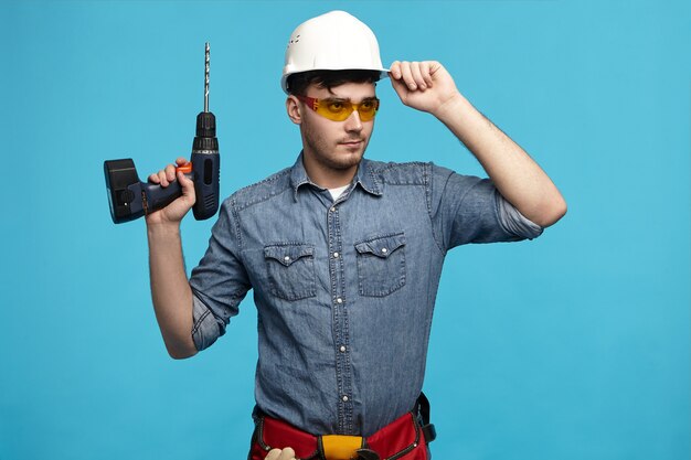 I'm ready to work. Horizontal shot of confident serious young unshaven male worker in safety eyewear holding drill