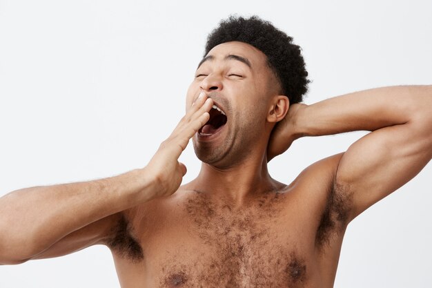 I want to sleep. Close up portrait of handsome athletic young afro american guy with curly hair and naked torso clothing mouth while yawning, going to bed after long day at work.