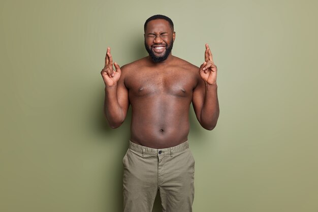 I want to win. Positive dark skinned African American man crosses fingers makes wish has hope in better poses with naked torso