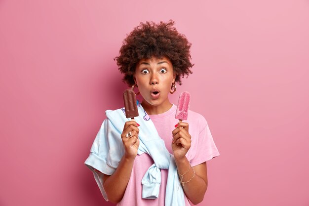 Ice cream time. Surprised dark skinned young woman poses with delicious frozen summer dessert stares bugged eyes dressed casually offers to taste it isolated over pink wall. Wow how yummy