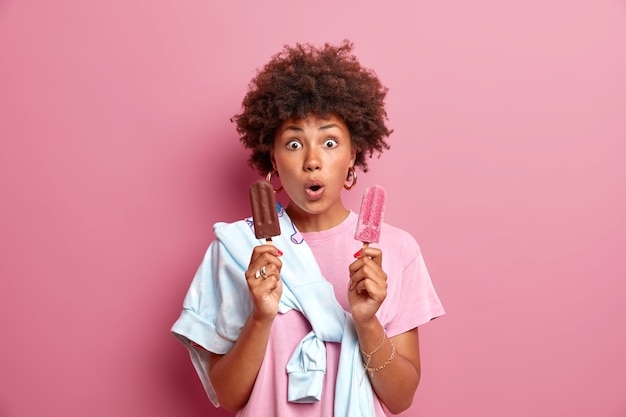 Бесплатное фото Время мороженого. удивленная смуглая молодая женщина позирует с восхитительным замороженным летним десертом, пристально смотрит на глаза, небрежно одетая предлагает попробовать его изолированно над розовой стеной. вау, как вкусно