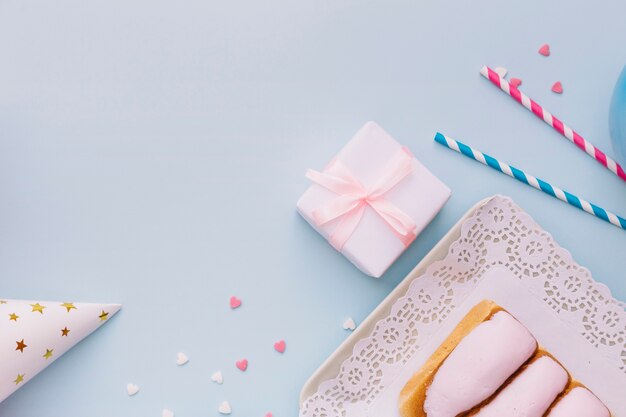 Iced buns; drinking straws; party hat; sprinkles and pink gift box on blue backdrop