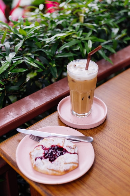 木製のテーブルの上のピンクのスタンドにガラスのアイスラテと夏のカフェの緑の茂みにクランベリーパイ