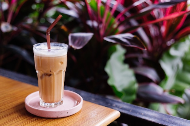 Iced latte in glass on a pink stand on wooden table in summer cafe green bushes