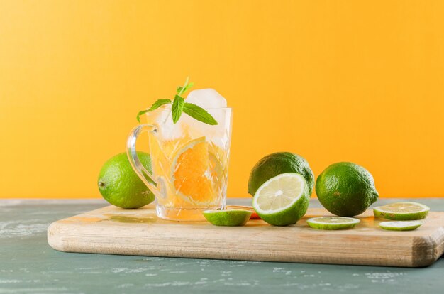 Icy detox water in a cup with orange, limes, mint, cutting board side view on plaster and yellow background