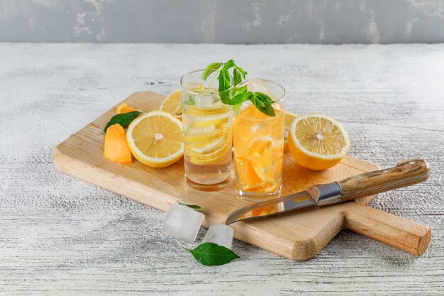 오렌지, 레몬, 민트, 나이프, 지저분한 및 석고 배경에 커팅 보드 높은 각도보기 유리에 얼음 해독 물