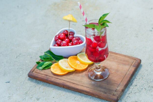 Ледяной напиток в стакане с вишней, лимоном, крупным планом листьев на цементе и разделочной доске