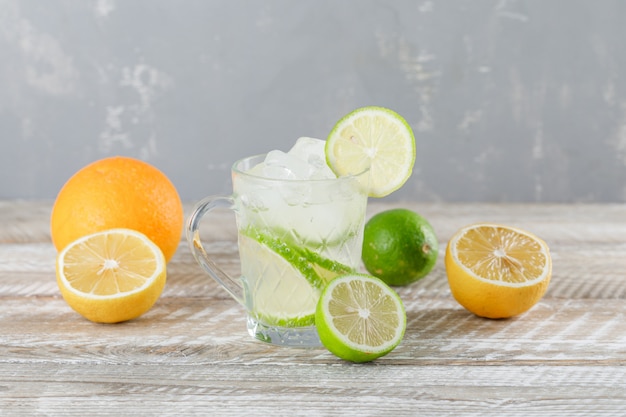 Ледяной мохито коктейль в чашке с лаймом, апельсином, лимоном сбоку на деревянной и гипсовой стене