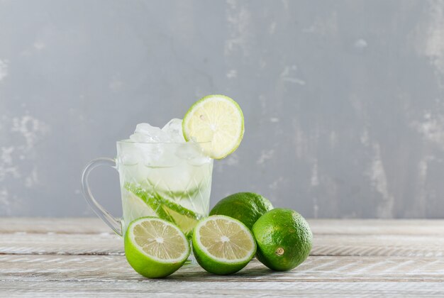 Icy mojito cocktail with limes in a glass cup on wooden and plaster background, side view.