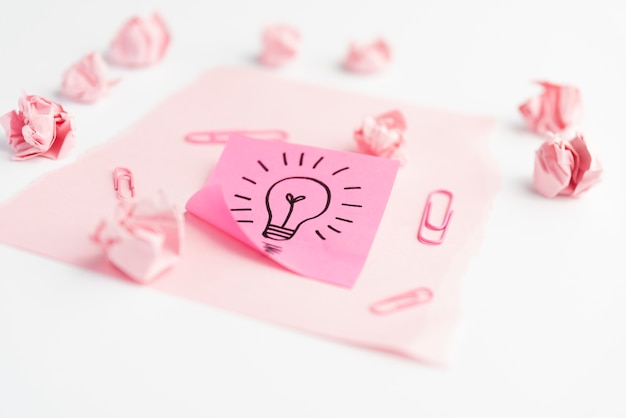 Idea icon on adhesive note with paperclip; crumpled paper and card paper over white desk