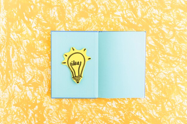 黄色のテクスチャ背景の上に青いページのノートブックのアイデア電球