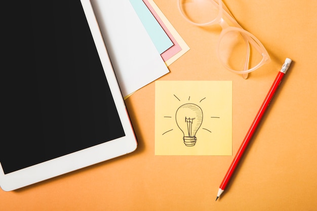 Idea symbol; pencil; eyeglasses; digital tablet and card paper on an orange backdrop