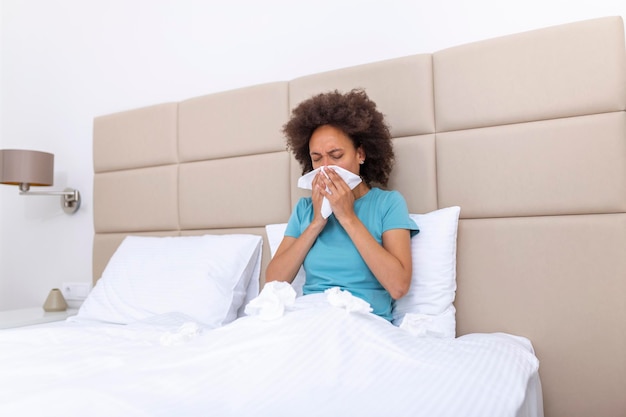 Больная африканская молодая женщина, покрытая одеялом, сморкается, заболела лихорадкой, простудилась, чихает в ткани, сидит на диване, больная аллергия, черная девушка с симптомами аллергии, кашляет дома концепция гриппа